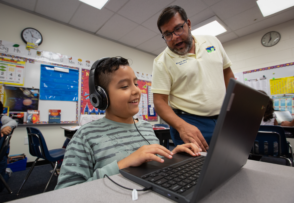 a young boy with headphones working on a computer gets help from his male teacher