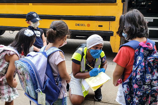 A school staff member writes down the name of a student, outside next to a school bus with other students nearby