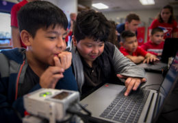 two young male students look at a computer and react to what they can make a small robot do