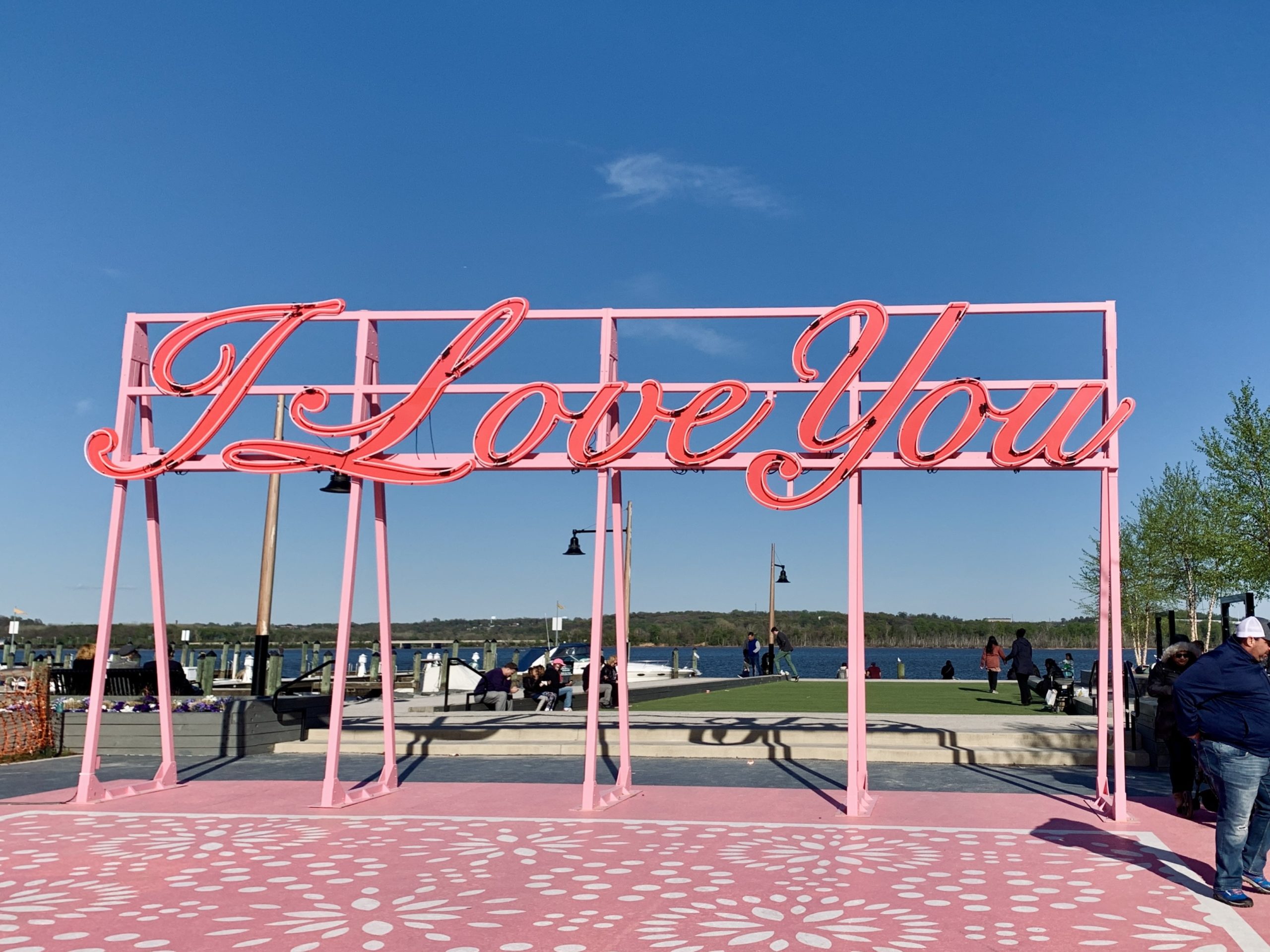 A pink, cursive I Love You sign sits in front of the blue sky. It is clear day on the Old Town Waterfront, and the rotating art installation is occupied.