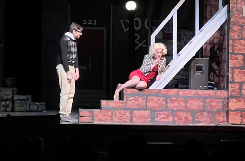 A young man in a sweater and slacks stands, acting, near steps, where a girl with strikingly blond hair and a red dress cowers hopelessly next to him. He tries to comfort her, but the distance is palpable. They are performing Little Shop of Horrors and the rest of the stage is dark.