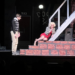 A young man in a sweater and slacks stands, acting, near steps, where a girl with strikingly blond hair and a red dress cowers hopelessly next to him. He tries to comfort her, but the distance is palpable. They are performing Little Shop of Horrors and the rest of the stage is dark.