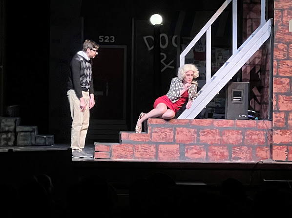 A young man in a sweater and slacks stands, acting, near steps, where a girl with strikingly blond hair and a red dress cowers hopelessly next to him. He tries to comfort her, but the distance is palpable. They are performing Little Shop of Horrors and the rest of the stage is dark.