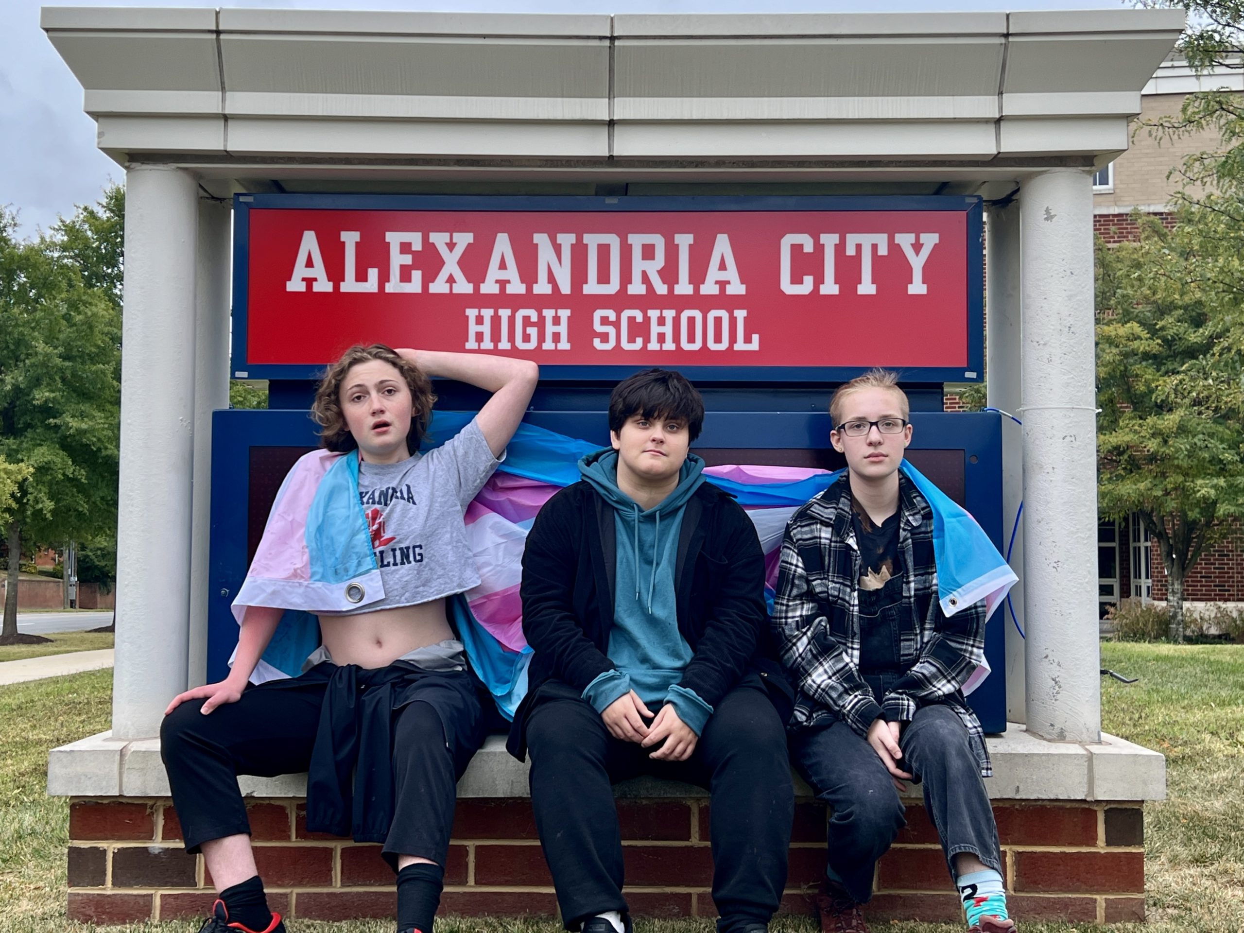 Three students sit at the sign for Alexandria City High School with a trans flag wrapped around their shoulders. The students are Chris, Michael Goldsberry, and KD Bectel.